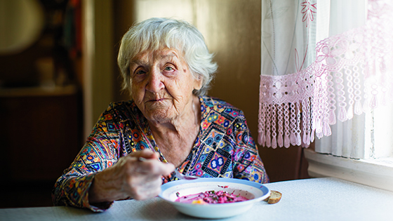 Ältere Frau sitzt am Tisch vor einem Teller Suppe