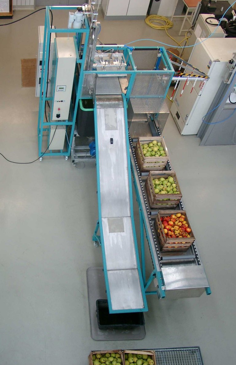Von oben fotografierte Anlage zur Heißwasserbehandlung von Bio-Äpfeln mit integriertem Laufband, auf dem Holzkisten mit unterschiedlichen Apfelsorten stehen